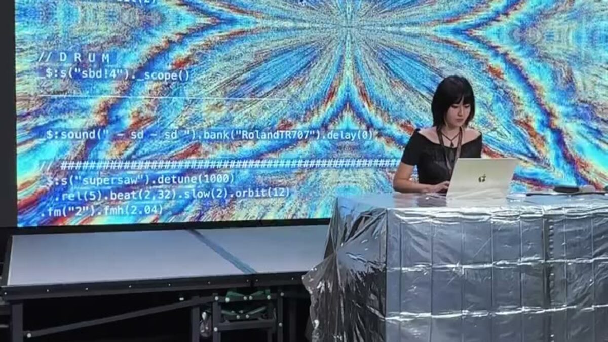 表演者在大型 LED 牆前操作筆電，牆上同時顯示程式碼與生成畫面